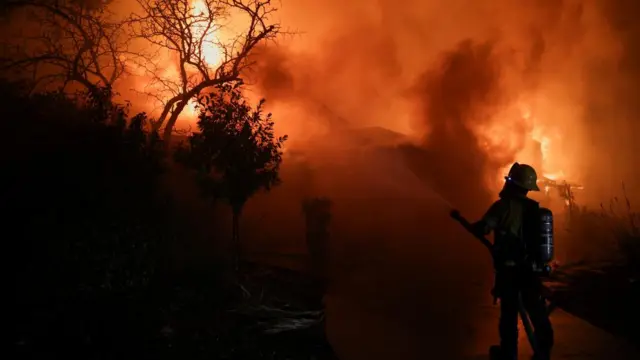 A firefighter works to extinguish flames in Pasadena, California. The sky is red, there are plumes of smoke and the shadow of trees.