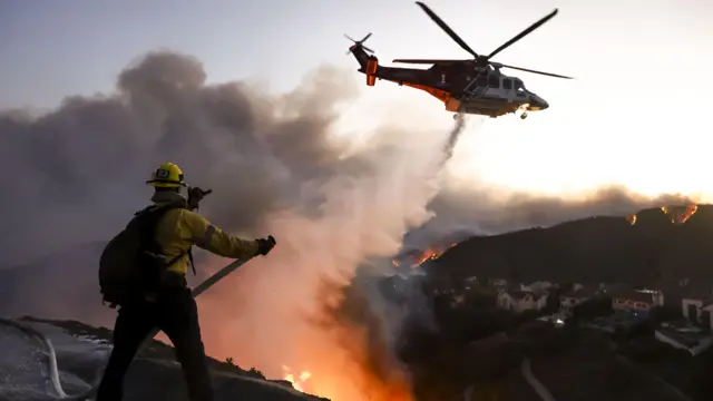 A firefighter uses a hose and water drops from a helicopter