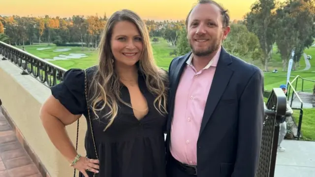 Miriam Feygenson, wearing a black dress, stands next to her husband Alex Filler, who wears a pink shirt and black suit. Behind them is a golf course.