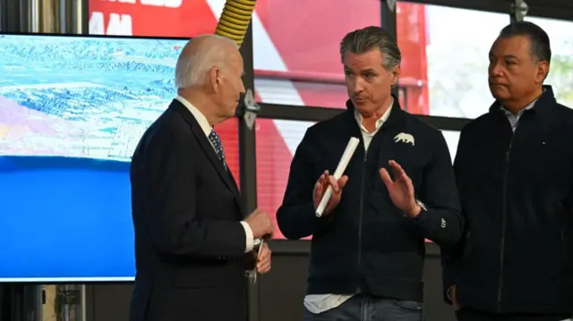 Gavin Newsom gestures with his hands as he explains something to President Joe Biden while the two men were briefed on the wildfires. A screen in the background shows the scale of the wildfires across the area