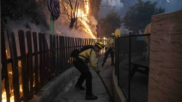 Two firefighters in gear hold a hosepipe as a fire rages on in the background.