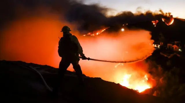 A Los Angeles County firefighter standing on a hilltop battles the Palisades wildfire.