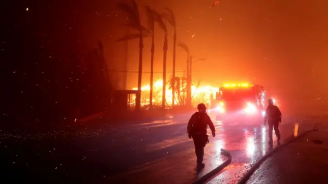 Fires rage near palm trees on a boulevard in Los Angeles. The silhouettes of firefighters can be seen against the headlights of an oncoming fire truck.