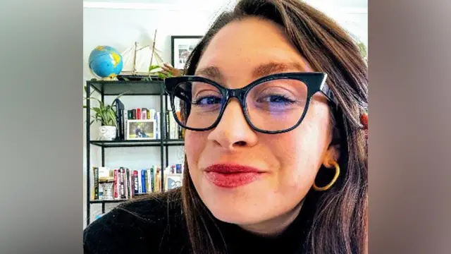 A woman with brown hair and brown eyes is wearing glasses and gold hoops as she poses for a picture.