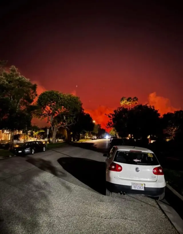 Red flames and smoke can be seen down a suburban tree-lined street.