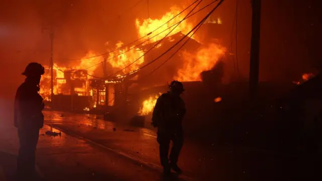 A fire rips through a structure and power lines in the background, with two firefighters in the foreground.