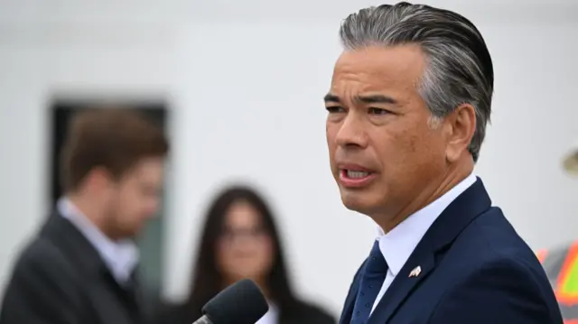 Rob Bonta wears a blue suit and tie speaking behind a podium with a microphone. Two people are in the background blurred out.