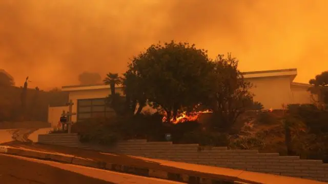 A brush fire burns near homes in Pacific Palisades, California. The sky is red and filled with smoke.