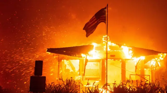 A U.S flag flies as fire engulfs a structure while the Palisades Fire burns during a windstorm on the west side of Los Angeles, California