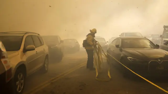 A Los Angeles firefighter drags a hose past cars trying to evacuate a neighborhood being threatened by the Palisades wildfire in Pacific Palisades, California