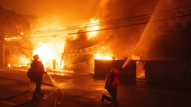 Two firefighters spraying water at a house that is being engulfed by flames