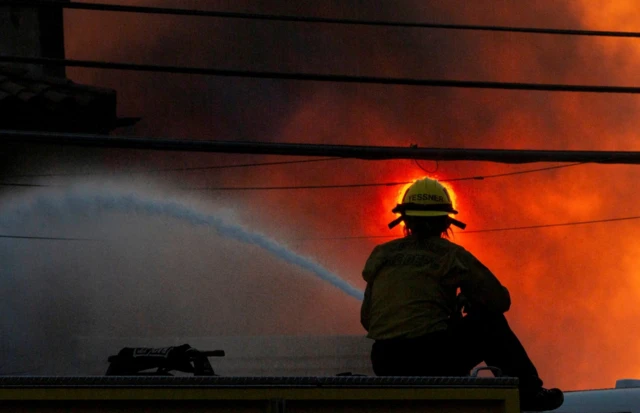 DA firefighter battles the Palisades Fire as it burns during a windstorm. The firefighter shoots water toward the blaze, the sky is filled with black and red smoke.