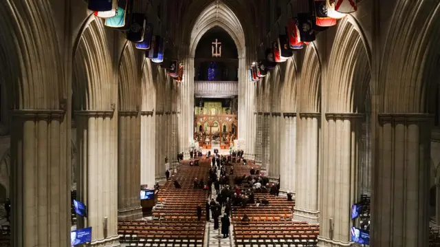 A long shot of the interior of the National Cathedral. Mourners are gathered in black in the middle