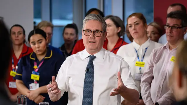 Starmer, with his jacket off and sleeves rolled up, stands in front of medical staff as he addresses a crowd