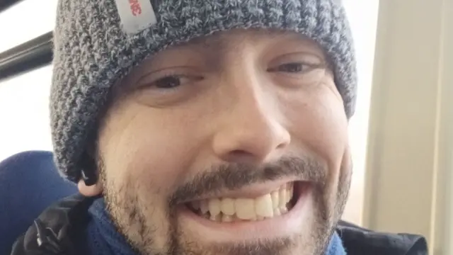 A man, Tyler Foyle, smiles to camera, wearing a hat and scarf