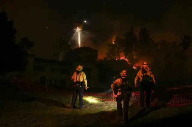 Night scene of three firefighters in a grassy field, stand facing fire in the background with a helicopter light shining down.