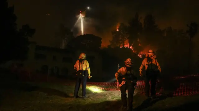 Three firefighters stand with protective equipment on and torches looking on at a fire blazing behind some trees. A helicopter with a light shining down is in the sky above it.