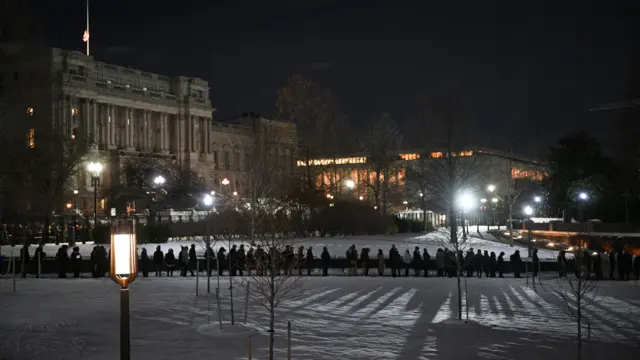 A distant view of a long line of mourners waiting in the snow at night.