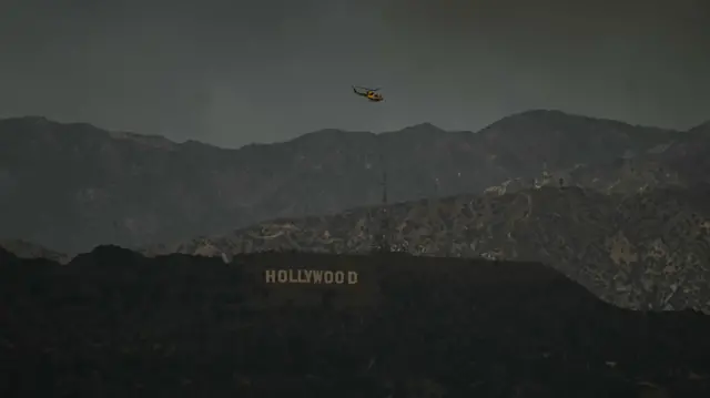 A firefighting helicopter flies above the Hollywood sign as the sky is engulfed in dark smoke from the many fires burning around the city.