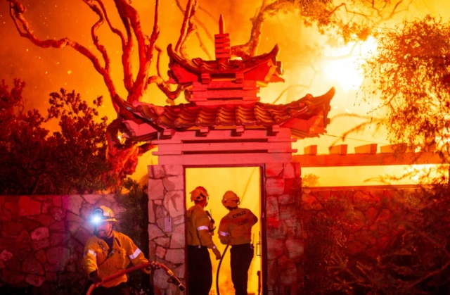 Firefighters battle the Palisades Fire as it burns during a windstorm on the west side of Los Angeles. The sky is entirely red with three firefighters holding a hose pipe, directly it toward the fire.