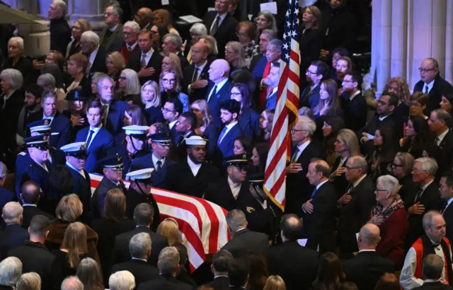 The casket covered by the US flag is brought into the cathedral as mourners look on