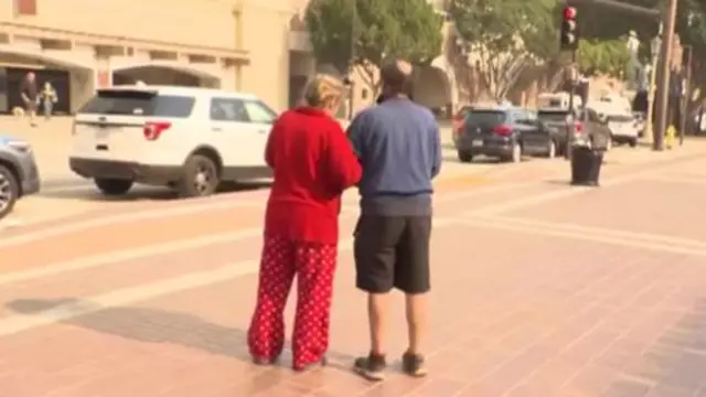 Un couple, bras croisés, vu de dos. L’un porte du rouge, l’autre un pull bleu et un short noir.