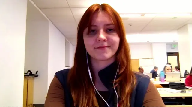 A woman on Zoom is talking to the camera. she has red hair and is wearing earphones with a cord.