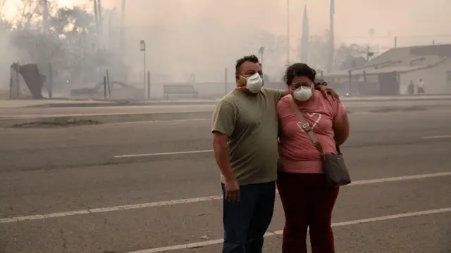 A man and woman react as they watch their home being affected by wildfire.