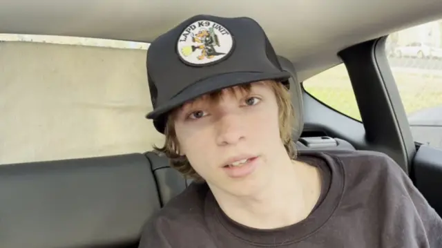 Arthur who lives near Eaton is sat in the back of a vehicle looking directly at the camera lens. He is wearing a back baseball cap and a black jumper