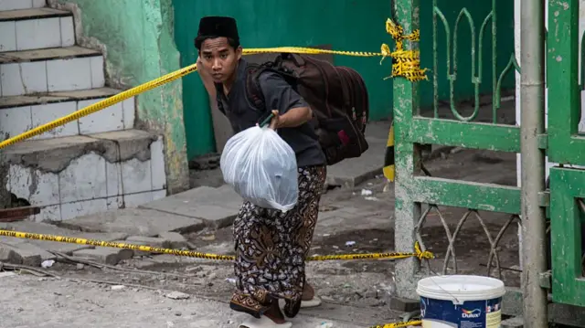A teenager in dark colour clothes carrying a backpack and white plastic bag bends down to cross a yellow cordon
