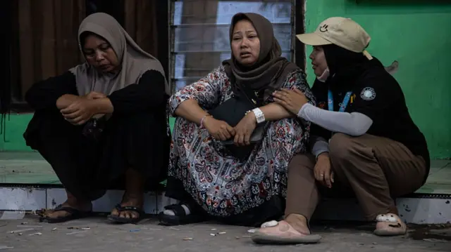 Three women sitting on a curb, with the woman in the middle looking distressed