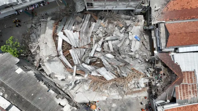 A photo shows the collapsed building from higher ground, with rubble in the center surrounded by other buildings.