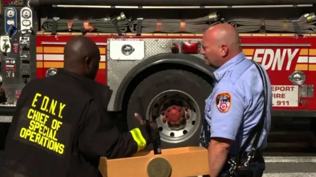 Two emergency responders stand near a podium with a microphone, with a fire truck in the background
