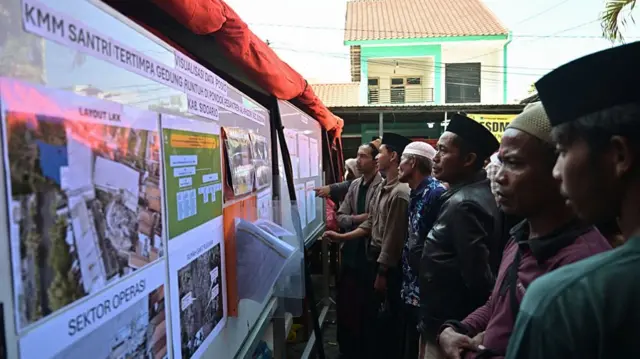 A row of people standing in front of a white board pasted with papers showing information about the collapsed building