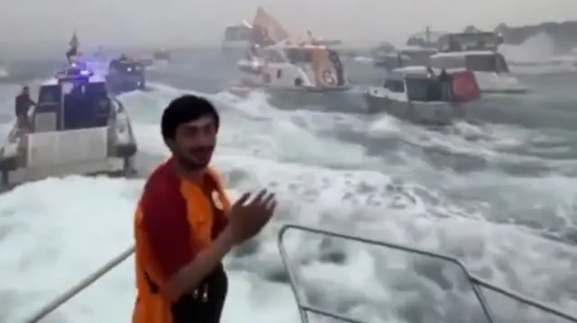 A man wearing a Galatasary shirt is clapping on the front of a boat. Half a dozen boats are visible, some waving Galatasary and Turkey flags.
