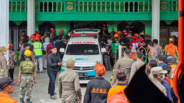 A crowd of rescuers in different colour uniforms gather around an ambulance parked at a green building
