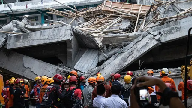 A crowd of rescuers with orange helmets stand in front of a collapsed building