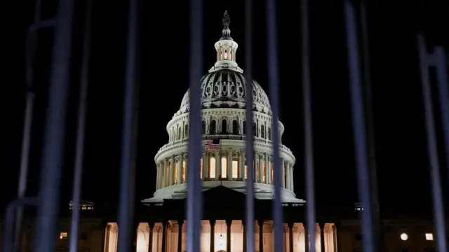 A white domed building can be seen through metal bars, lit by floodlights. The Stars and Stripes can be seen flying just beneath the dome.