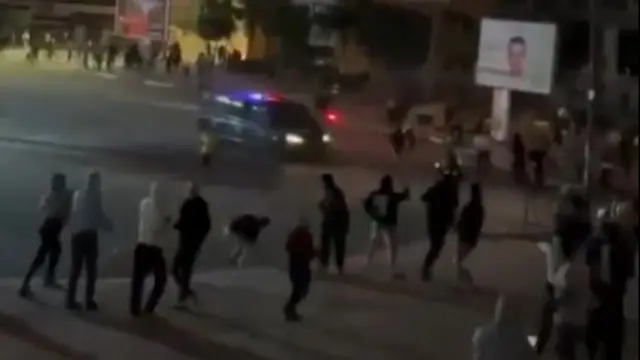 A screenshot of a police van moments before it hits a protester. Other people can be seen in the foreground looking on