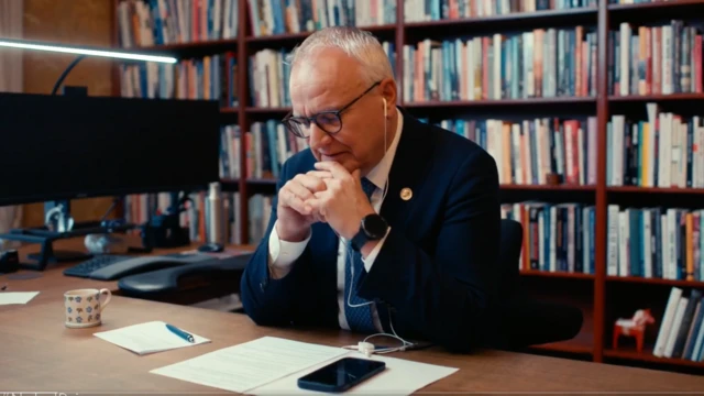 A man in a dark suit in a study is seen making a phone call via wired headphones on his mobile device