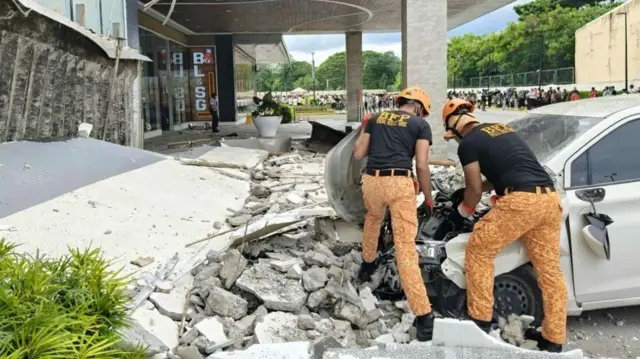 Two firemen inspect a crushed hood of a car, standing on a pile of rubble