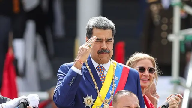 Maduro saluting as he wears a large ornamental gold chain, a badge and a large gold ring. He has a sash on and a woman smiles behind him