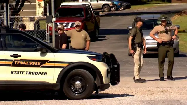 Three police officers stand outside the gate to the the Accurate Energetic Systems military explosives plant. They stand behind a police car which reads Tennessee state trooper on the door.