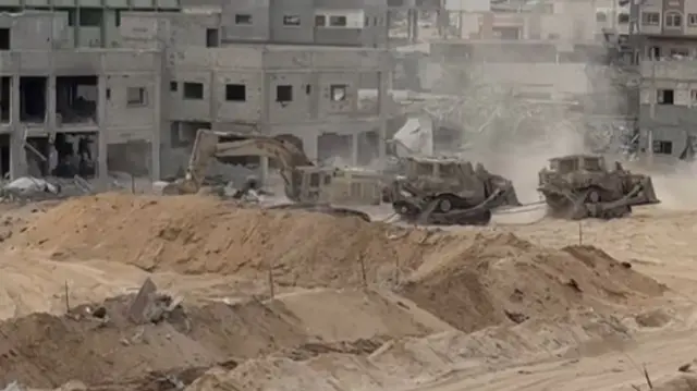 A bulldozer and two military vehicles moving in the midst of rubble.