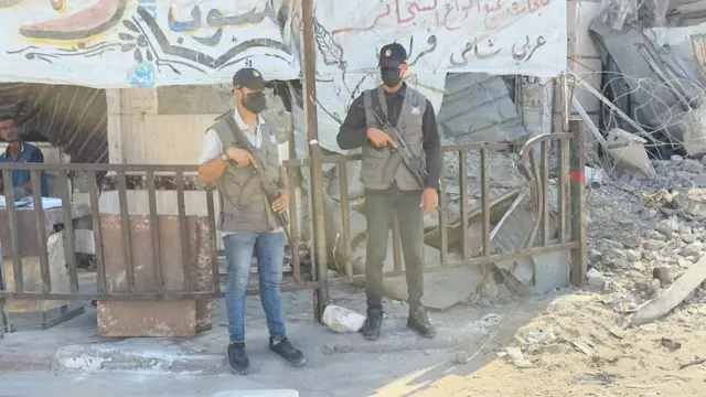 Two Hamas gunmen stand holding guns in front of a temporary shelter