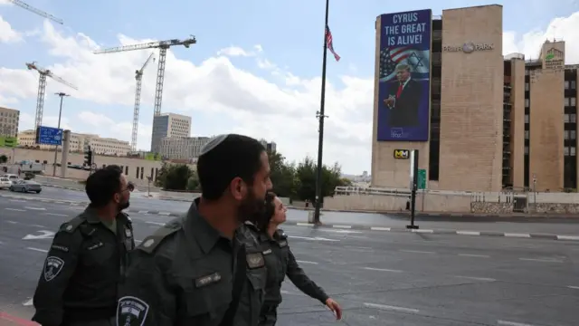 Three police officers walk past a billboard reading "Cyrus The Great Is Alive!" with Donald Trump pictured