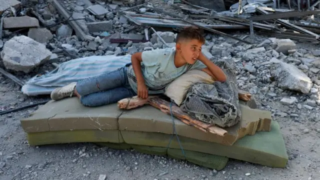 A boy lies on top of a bundle of mattresses in amongst rubble
