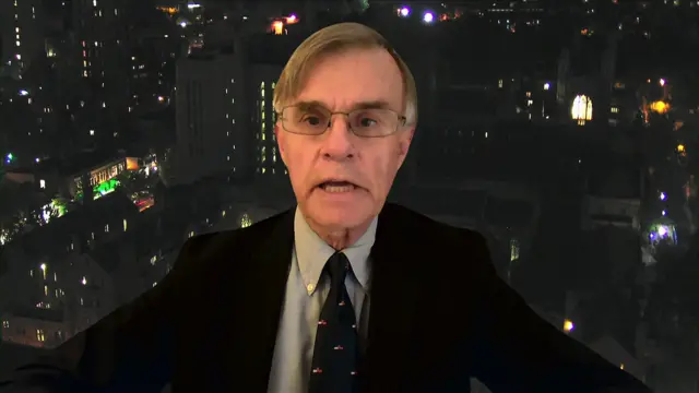 Gordon Gray, wearing a dark suit and tie, and glasses, speaking during an interview. The background is of a evening cityscape of buildings