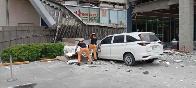 Backview of two firemen standing near a white car. In front of them is a broken building structure