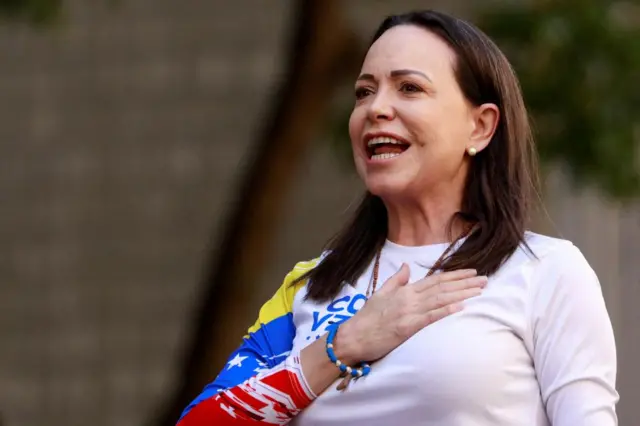 Maria Corina Machado in white top with colours of her nation's flag, holding her right hand to her chest